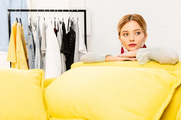 Pensive girl in earrings sitting on yellow sofa at home — Stock Photo