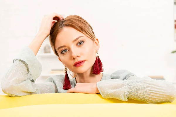 Pensive girl in earrings sitting on yellow sofa at home — Stock Photo
