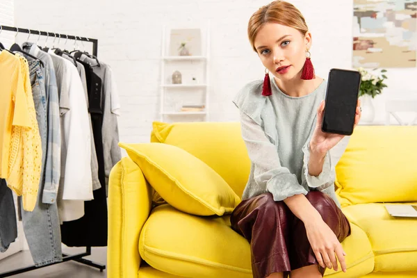 Очаровательная девушка в модных серьгах сидит на желтом диване и использует смартфон дома — стоковое фото
