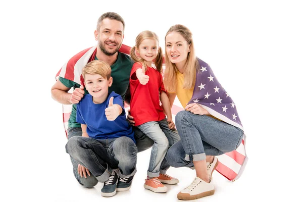 Familia sonriente con bandera americana mirando a la cámara y mostrando los pulgares hacia arriba aislados en blanco - foto de stock