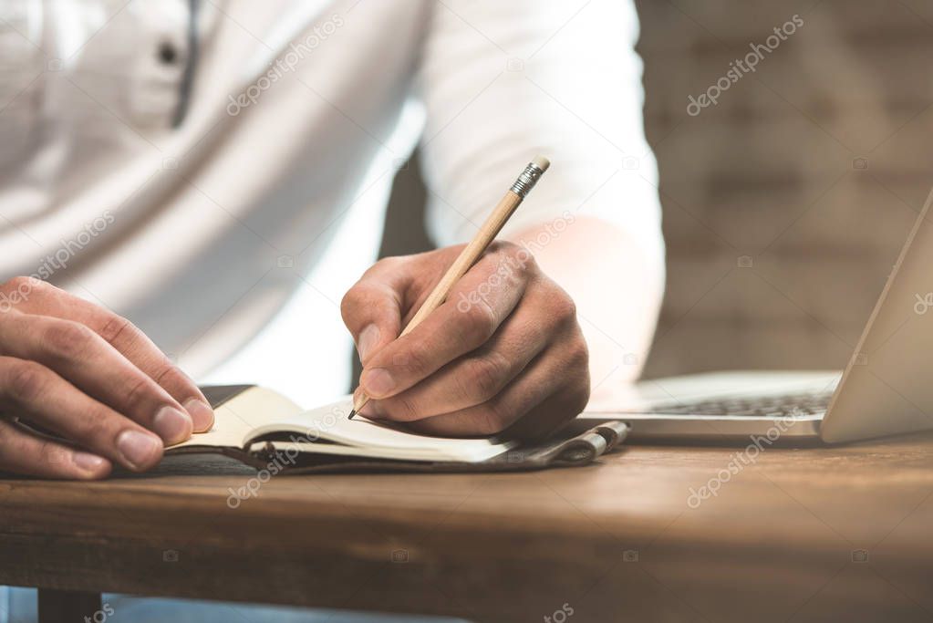 partial view of man making notes in textbook at table with laptop