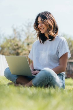 Laptop kullanan ve yeşil çimlerin üzerinde oturan mutlu bağımsızların seçici odak noktası 