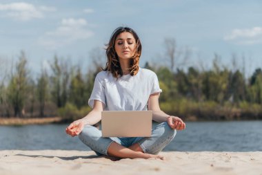 Gözü kapalı çekici bir serbest yazar dizüstü bilgisayarın yanında kumlara meditasyon yapıyor. 