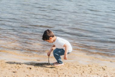Çıplak ayaklı çocuk ıslak kum ve göletin yanında sopa tutuyor. 