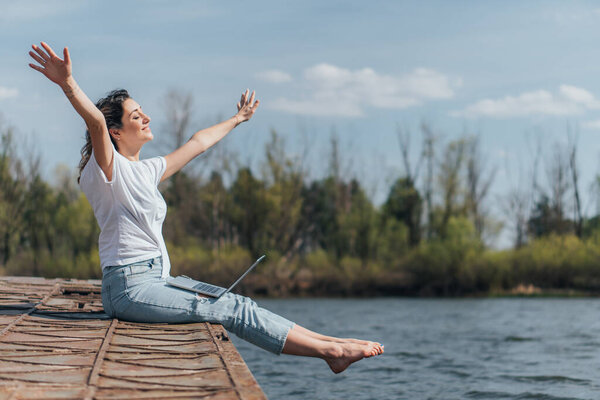 cheerful woman with outstretched hands sitting near lake 