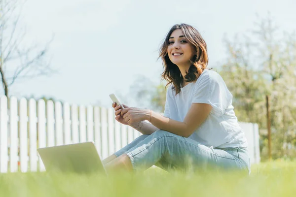 Selektywna Ostrość Radosna Kobieta Trzyma Smartfona Pobliżu Laptopa Siedzi Trawie — Zdjęcie stockowe