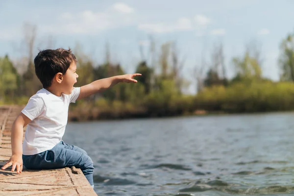 川の近くに座っている間に指で指差す感情的な少年のプロフィール — ストック写真