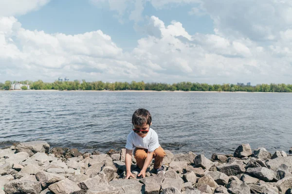Anak Bergaya Berkacamata Hitam Menyentuh Batu Ketika Duduk Dekat Danau Stok Gambar