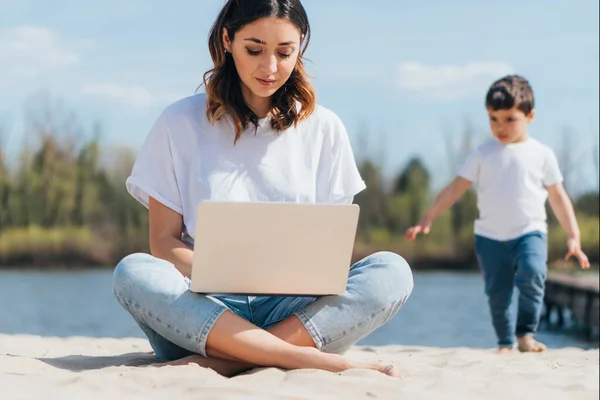 Foco seletivo de freelancer usando laptop enquanto sentado na areia perto de filho bonito — Fotografia de Stock