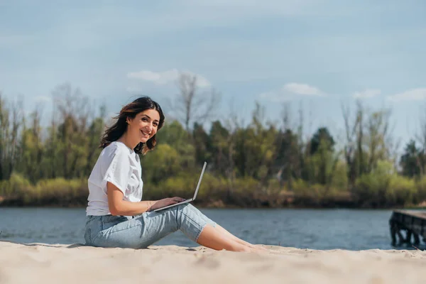 Вибірковий фокус щасливого фрілансера за допомогою ноутбука біля озера — стокове фото