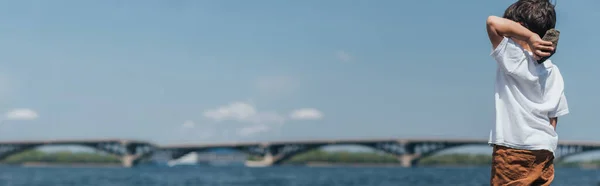 Panoramic crop of boy holding rock near blue lake — Stock Photo