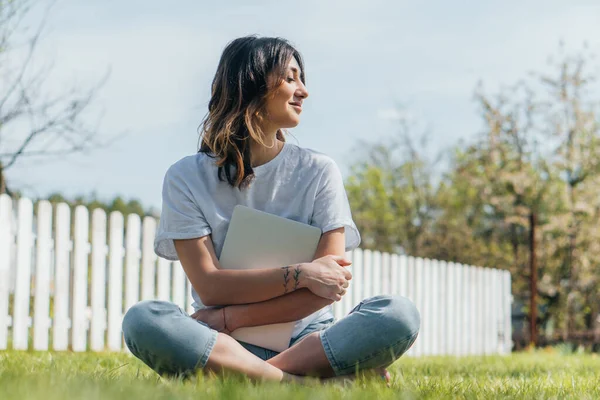 Избирательный фокус веселой женщины, сидящей на траве и держащей ноутбук — стоковое фото