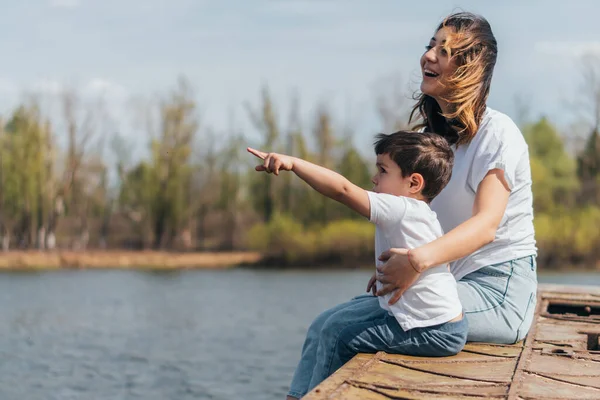 Mignon enfant pointant du doigt tout en étant assis près de mère heureuse et le lac — Photo de stock