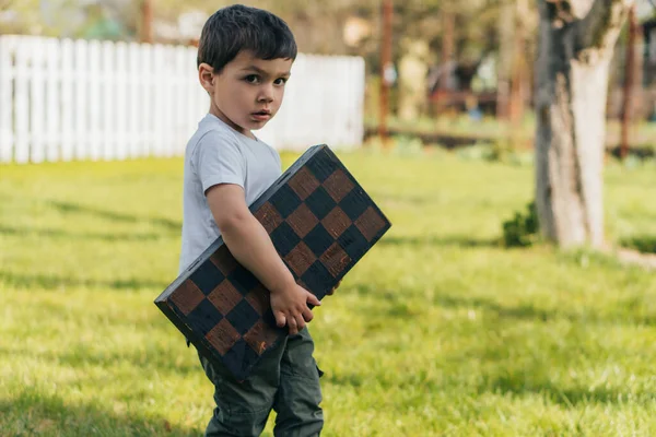Милый ребенок держит шахматную доску и смотрит в камеру — стоковое фото