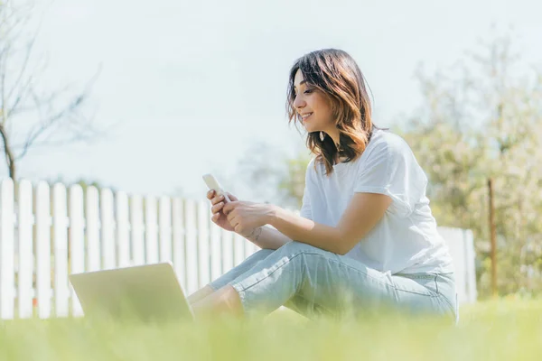 Foco seletivo da mulher feliz usando smartphone perto do laptop enquanto sentado na grama — Fotografia de Stock