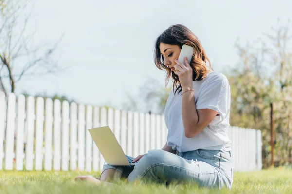 Foyer sélectif de femme heureuse parlant sur smartphone et en utilisant un ordinateur portable tout en étant assis sur l'herbe — Photo de stock