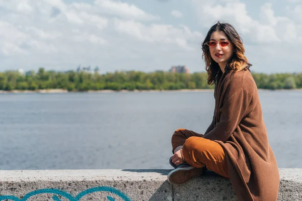 Stylish woman in sunglasses sitting near river and looking at camera — Stock Photo