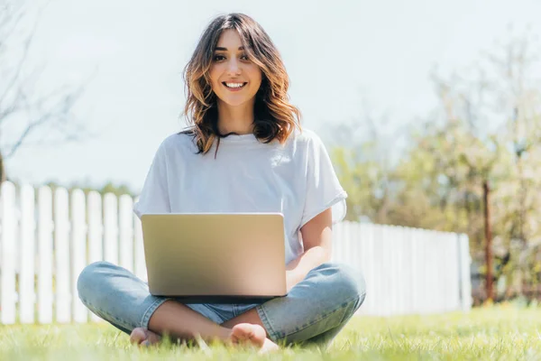 Selective focus of happy freelancer sitting with laptop on grass — Stock Photo