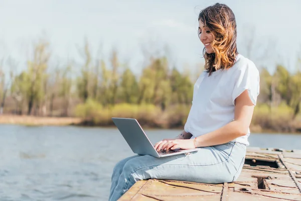 Feliz freelancer sonriendo y utilizando el ordenador portátil cerca del río - foto de stock