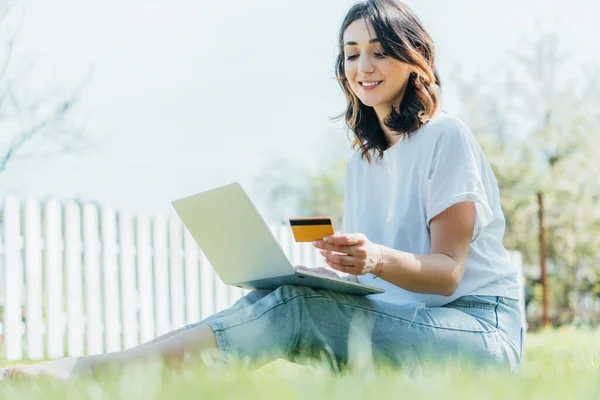 Foyer sélectif de femme heureuse regardant la carte de crédit près de l'ordinateur portable et assis sur l'herbe — Photo de stock