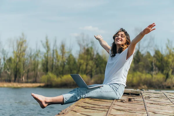Весела жінка з витягнутими руками сидить з ноутбуком біля озера — стокове фото