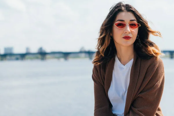 Trendy woman in stylish sunglasses looking at camera near lake — Stock Photo