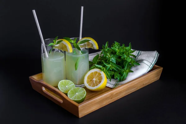Homemade citrus lemonade in glasses with straws with mint leaves and a bunch of mint, lemons, limes on a bamboo tray and whte towel on a black background. Front view