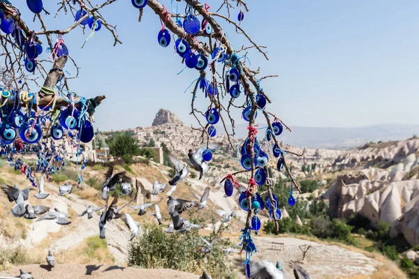 Evil Eye Beads on Tree and Fairy tale chimneys on background of blue sky in Guvercinlik Valley,Goreme,Turkey.Branches of the old tree decorated with the eye-shaped amulets,Nazars,Evil eye,made of blue glass.
