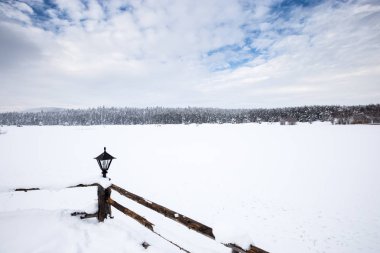 Bulutlu bir sıkıcı gün çam ormanı ile kış manzara görünümü. Çit ve ön plan üzerinde küçük bir lamba.