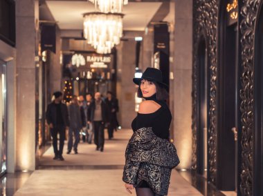 Siyah koridorda gülümseyen şapkalı güzel bir manken. Otel koridorunda poz veren klasik elbiseli seksi bir kadının portresi.