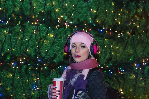 Beautiful woman in beanie hat with takeaway coffee and headphones poses in front of Christmas decoration lights.