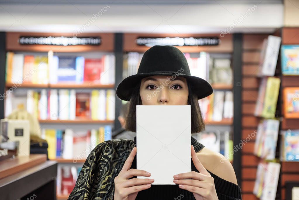 Attractive beautiful woman with black hat holding book partially covered face with closed book and looking at camera.