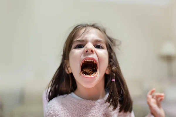 Little cute girl opens her big mouth and shows chewed eats chocolate with funny face while looking at camer