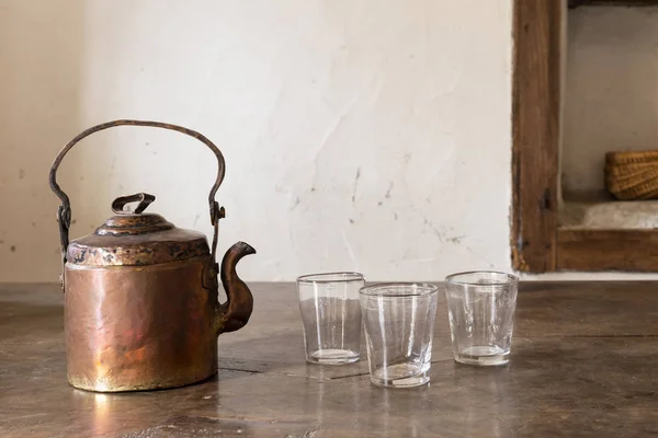 Still life with a beautiful worn retro style copper tea kettle with three empty glasses on a massive wooden table.