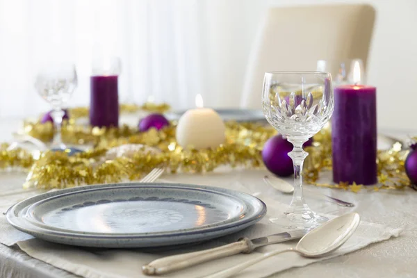 Close-up of a decorated dining table with burning candles, sterling cutlery, crystal glasses, colorful christmas balls and gold tinsel