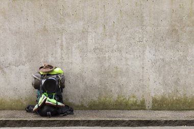 Sırt çantası ile yeşil havlu, kızartma tavası, uyku tulumu, deri şapka ve yeşil koşu ayakkabıları üzerinde gri bir duvar önünde yol kenarında duran hiking
