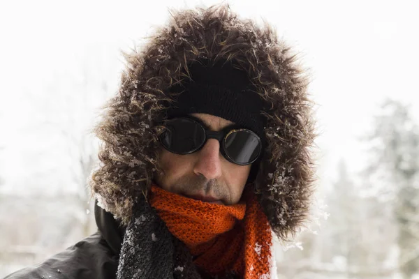 Portrait of a single male winter adventurer wearing a warm green coat with fur hood, a blue ski cap, an orange scarf and black retro style goggles