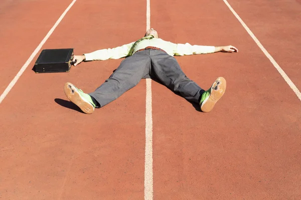 An exhausted business man in gray suit with green shirt and tie, black briefcase and broken green running shoes lying on a running track.