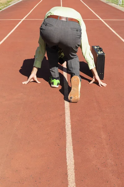 A business man in gray suit with green shirt, black briefcase and broken green sports shoes getting ready on a running track.