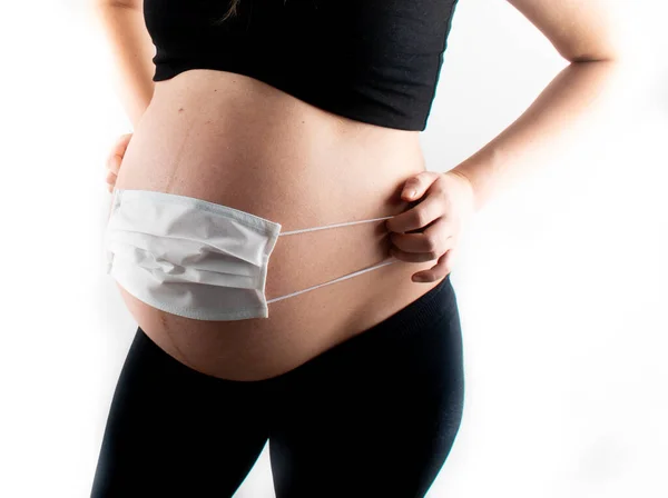 Pregnant woman holding white medicinal face mask on her pregnant belly abdomen. Coronavirus covid19 new normal rules and regulations, isolation and new lifestyle concept