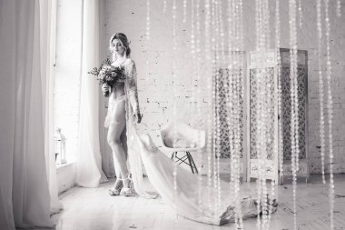 Gelin, sabahlığı ve iç çamaşırı giymiş pencere duruyor ve gelinlik onun elinde tutar. Sabah gelinin. Siyah beyaz fotoğraf.