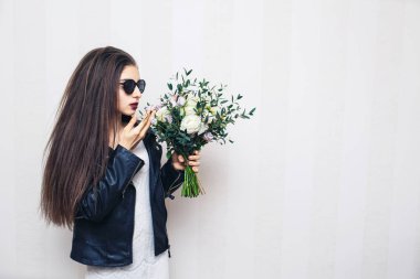 Güneş gözlüğü, kız güzel şık giyinmiş deri ceketi, buket çiçek ve beyaz duvara poz holding