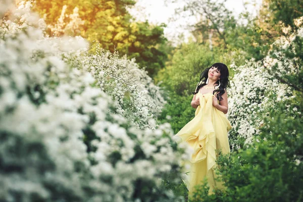 Chica feliz en vestido amarillo sonríe en el parque al atardecer entre arbusto de espirea . — Foto de Stock