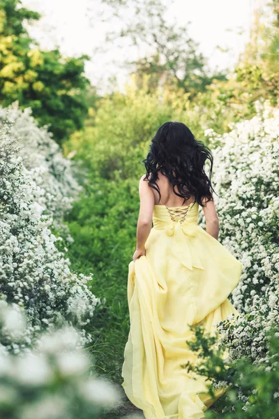 Menina em vestido amarelo corre ao longo do caminho entre arbustos floridos de spiraea — Fotografia de Stock