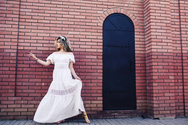 Hermosa chica en vestido blanco está de pie cerca de la pared de ladrillo con puerta de metal y posando — Foto de Stock