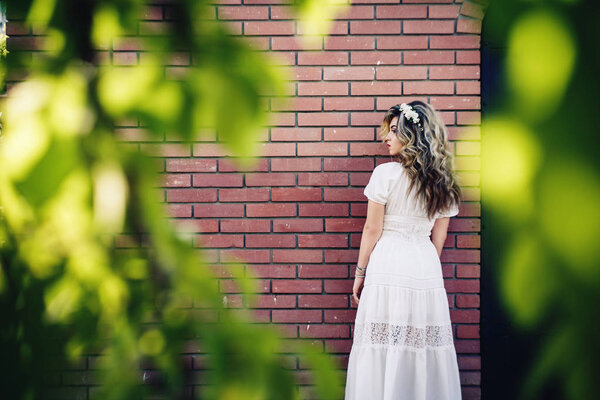 Beautiful girl in white dress is standing near brick wall with metal door and posing. Frame made of green leaves.