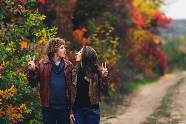 Coppia allegra mostra emozioni. uomo e donna in giacche di pelle e jeans si mostrano lingue sullo sfondo degli alberi autunnali . — Foto Stock