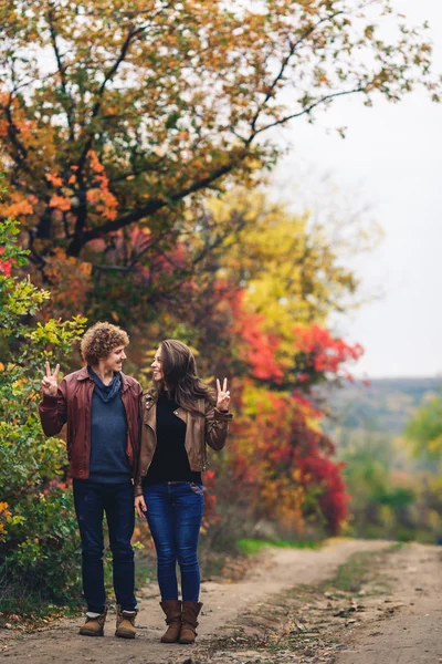 Pareja alegre muestra emociones. hombre y mujer en chaquetas de cuero y pantalones vaqueros contra el fondo de los árboles de otoño . — Foto de Stock