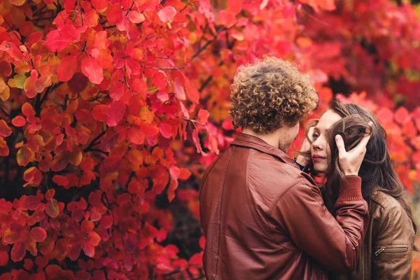 Curly-haired besnorde man en bruinharige vrouw knuffelen in de herfst tegen de achtergrond van rode bomen. — Stockfoto