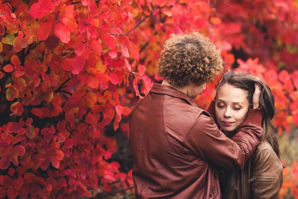 Curly-haired mustachioed man and brown-haired woman hugging in autumn against background of red trees. — Stock Photo, Image